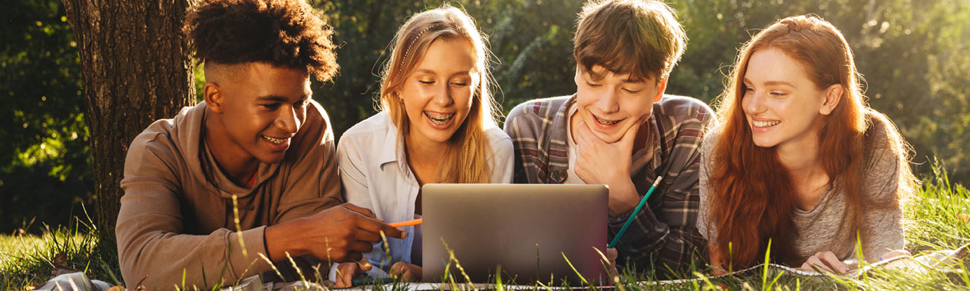 Group of smiling multhiethnic students doing homework together at the park, using laptop computer and mobile phone, laying on a grass